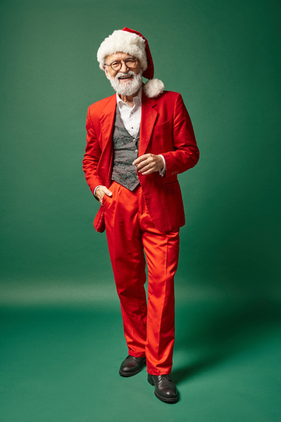 fröhlicher Mann als Weihnachtsmann gekleidet mit weihnachtlichem Hut posiert mit einer Hand in der Tasche, winterliches Konzept - Foto, Bild