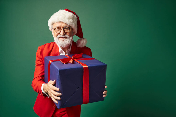 handsome stylish man in christmassy red hat and glasses holding huge blue present, winter concept - Foto, imagen