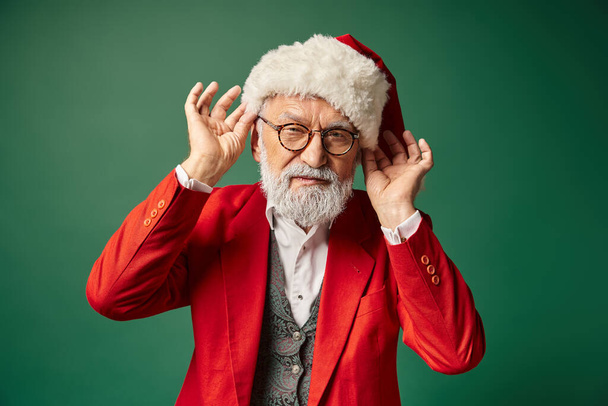handsome elegant Santa with white beard looking at camera touching hat with hands, winter concept - Photo, Image