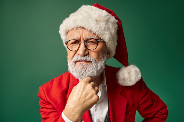 homme sérieux habillé en Père Noël regardant directement la caméra avec la main sous le menton, concept d'hiver - Photo, image