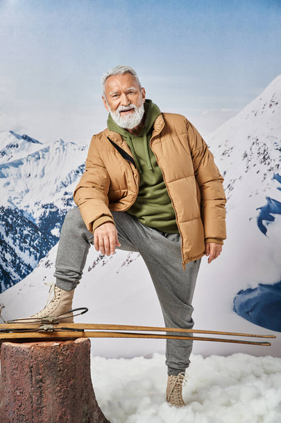 sportieve kerstman poseren met opgeheven been in de buurt van ski 's met besneeuwde berg op de achtergrond, winter concept - Foto, afbeelding
