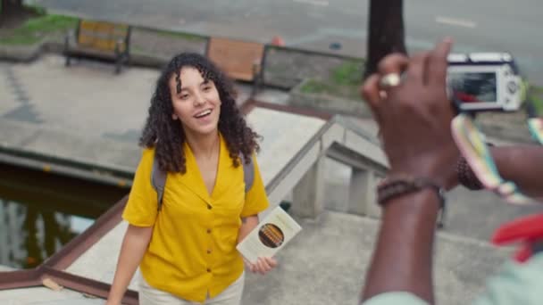 Среднеугловой снимок улыбающейся женщины с кудрявыми волосами, рюкзаком и туристической брошюрой, позирующей на пешеходном мосту через оживленную улицу, и неузнаваемым темнокожим парнем, фотографирующим - Кадры, видео