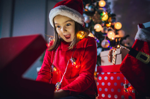 La niña está leyendo un libro sobre Navidad. Luces coloridas, sombrero de Papá Noel y árbol de Navidad - Foto, imagen