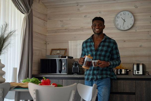 Afro-Amerikaanse man zit in een gezellige keuken, hij schenkt thee in een kopje - Foto, afbeelding