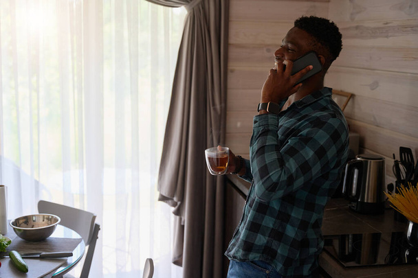 A fickó online kommunikál a mobilján, leül egy csésze teával a konyhában. - Fotó, kép