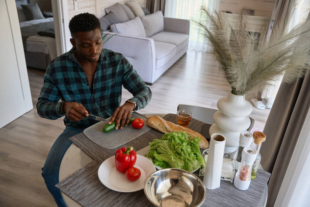 Cute African American facet przygotowuje sałatkę z soczystych warzyw, on jest w komfortowych ubrań casual - Zdjęcie, obraz