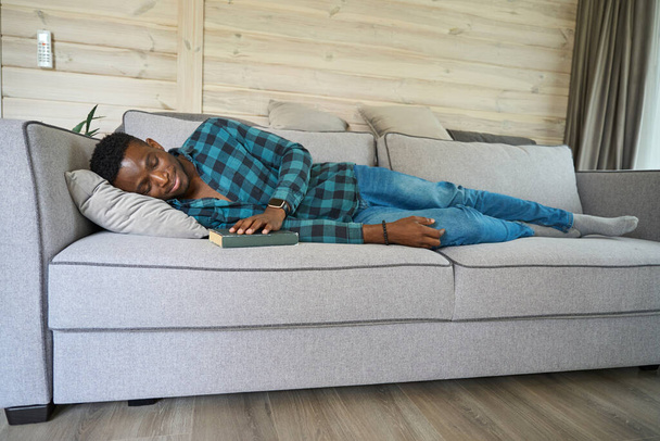 Afroameričan usnul s knihou na pohovce, na sobě měl kostkovanou košili a džíny. - Fotografie, Obrázek