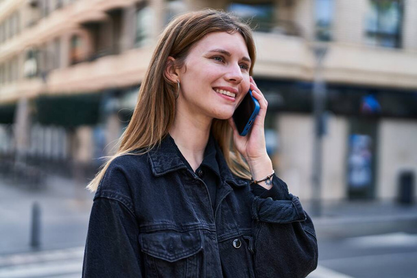 Молодая блондинка улыбается уверенно разговаривая по смартфону на улице - Фото, изображение