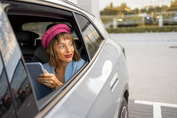 Porträt einer jungen, stylischen Frau, die mit Handy auf dem Rücksitz eines Autos sitzt, Blick aus dem Fahrzeugfenster. Frau mit rosa Hut, mit Gadget unterwegs - Foto, Bild