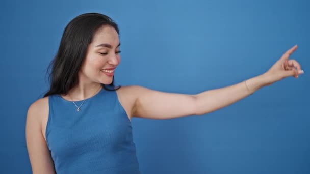 Nuori kaunis latino nainen hymyilee osoittaa sivulle sormet yli eristetty sininen tausta - Materiaali, video