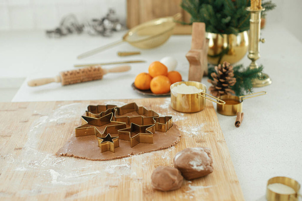 Masa de pan de jengibre con cortadores de metal dorado festivos en tablero de madera con harina, especias de cocina, decoraciones festivas en la cocina blanca moderna. Hacer galletas de jengibre de Navidad - Foto, imagen