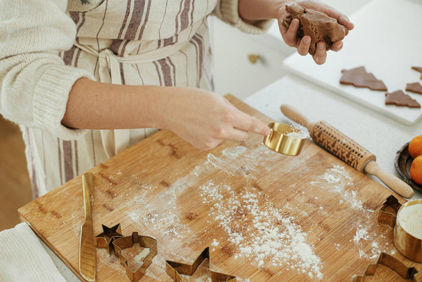 Χέρια ζυμώνοντας ζύμη μελόψωμο σε ξύλινη σανίδα με πλάστη, χρυσά μεταλλικά κόπτες, μπαχαρικά μαγείρεμα, εορταστική διακόσμηση στη σύγχρονη λευκή κουζίνα. Γυναίκα που φτιάχνει χριστουγεννιάτικα μπισκότα με μελόψωμο - Φωτογραφία, εικόνα