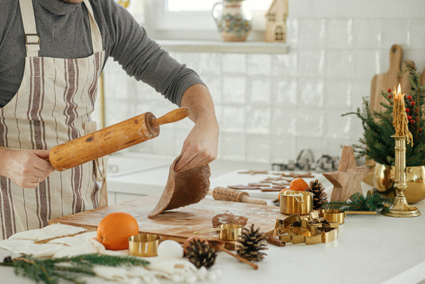 Ο άνθρωπος κάνει χριστουγεννιάτικα μπισκότα μελόψωμο στη σύγχρονη λευκή κουζίνα. Χέρι ζυμώνοντας ζύμη μελόψωμο σε ξύλινη σανίδα με αλεύρι, πλάστη, χρυσό κόφτη μετάλλων, το μαγείρεμα μπαχαρικά στον πάγκο - Φωτογραφία, εικόνα