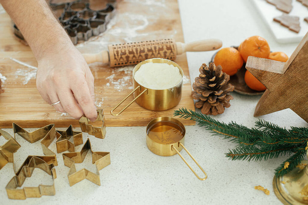 Hands cutting gingerbread dough with festive golden metal cutters on wooden board with flour, cooking spices on countertop in modern white kitchen. Man making christmas gingerbread cookies - Photo, Image