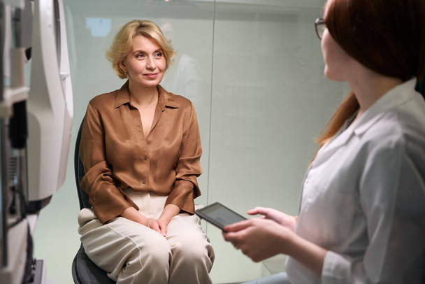 Keski-ikäinen nainen potilas silmälääkärin vastaanotolla, naiset kommunikoivat valoisassa huoneessa - Valokuva, kuva