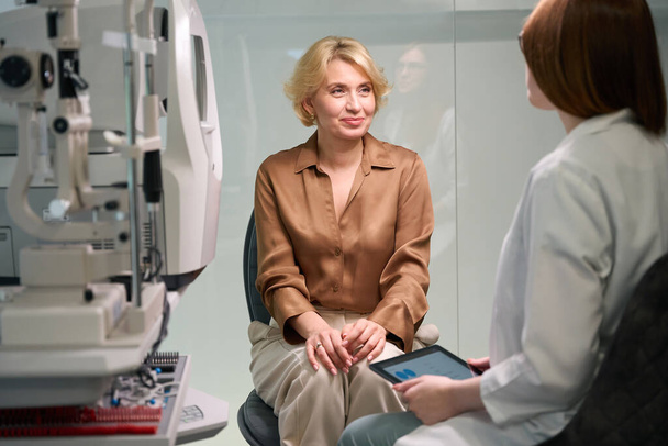 Κοκκινομάλλης οφθαλμίατρος συμβουλεύεται μια μεσήλικη κυρία σε μια δεξίωση, υπάρχουν σύγχρονες επαγγελματικές συσκευές στο δωμάτιο - Φωτογραφία, εικόνα