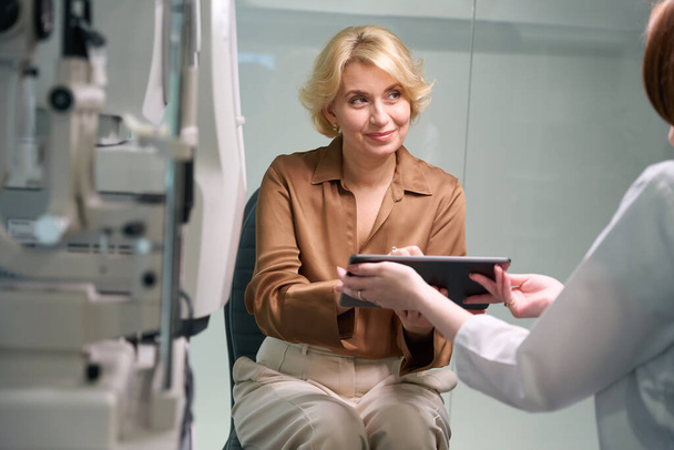 La oftalmóloga consulta a una señora de mediana edad, la paciente firma un contrato - Foto, imagen