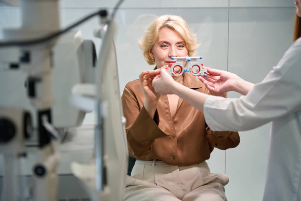 Η ασθενής με καφέ μπλούζα φοράει ειδική συσκευή για τον έλεγχο της όρασης, δίπλα σε ένα νεαρό γιατρό. - Φωτογραφία, εικόνα