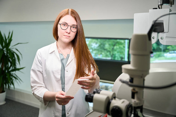 Pleasant young ophthalmologist at her workplace, professional devices indoors - Photo, Image