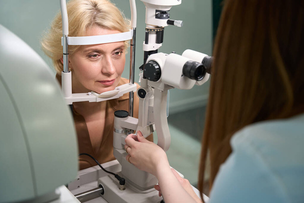 Μεσήλικη γυναίκα σε ραντεβού με οφθαλμίατρους, ο γιατρός χρησιμοποιεί μια σύγχρονη συσκευή στην εργασία - Φωτογραφία, εικόνα