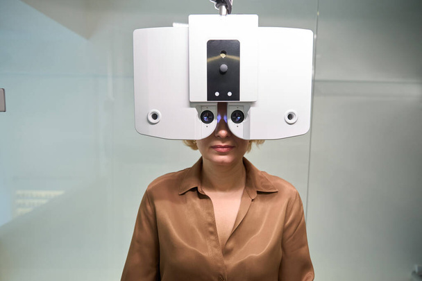 Dame en chemisier brun subit un examen ophtalmologique, un dispositif moderne est utilisé à un rendez-vous avec un ophtalmologiste - Photo, image