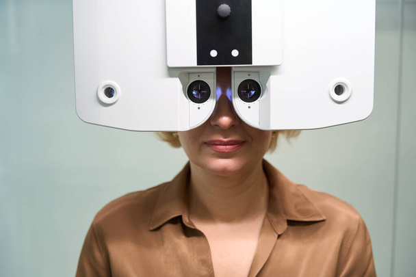 Женщина в коричневой блузке проходит офтальмологическое обследование, на приеме у офтальмолога используется современное устройство - Фото, изображение