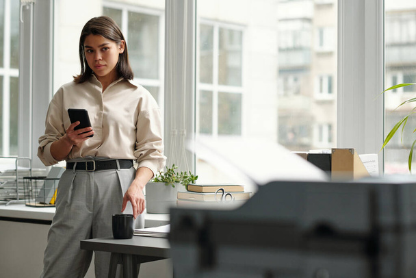 Nuori sihteeri, jolla on tummat hiukset, katselee asiakirjoja tulostimessa ja käyttää älypuhelinta seisoessaan tilavan toimiston ikkunaa vasten - Valokuva, kuva