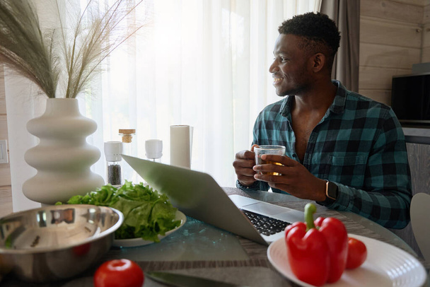 African American man sits in a cozy kitchen with a laptop, juicy vegetables for salad on the table - Photo, Image