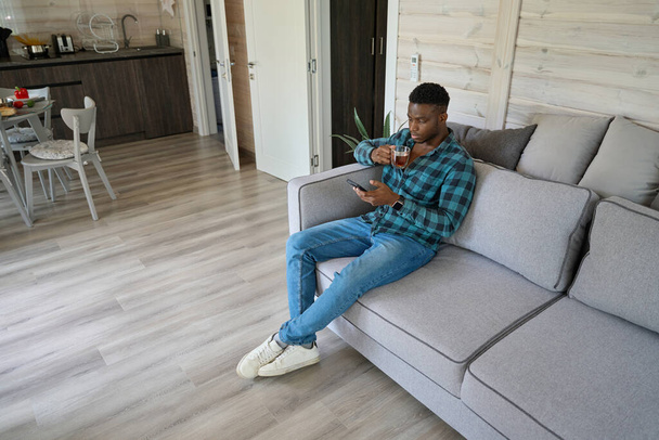 Afroamerykanin z filiżanką herbaty i telefonu komórkowego siedzi na kanapie, znajduje się w jasnym pokoju - Zdjęcie, obraz