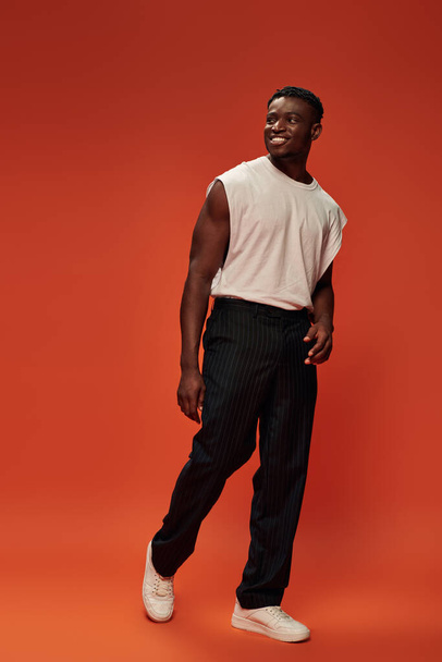 hombre americano africano alegre y caliente en camiseta blanca y pantalones negros mirando hacia otro lado en el fondo rojo - Foto, imagen