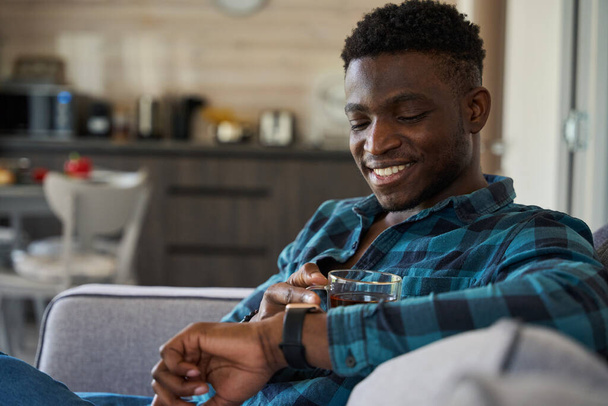 Afrikkalainen amerikkalainen mies katsoo älykästä kelloa kädessään, hän istuu sohvalla kupin teetä kanssa. - Valokuva, kuva