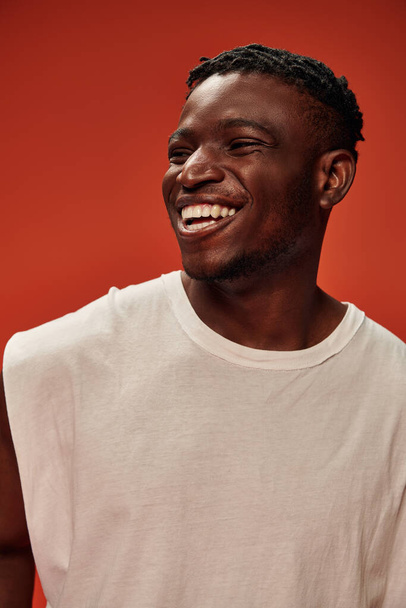 aufgeregter afrikanisch-amerikanischer Mann in weißem Tank-Top, lachend und wegschauend auf rotem Hintergrund, Glück - Foto, Bild