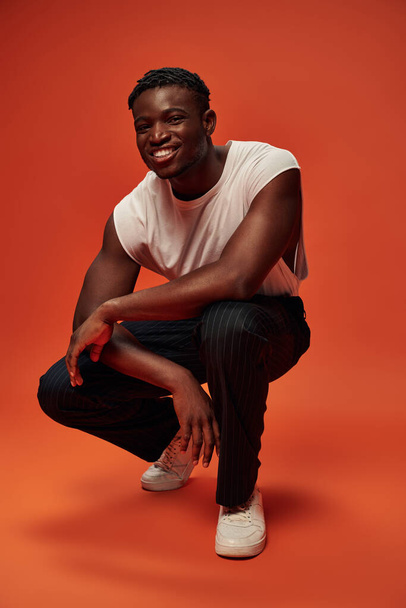 carefree african american man in trendy street wear sitting on haunches on red and orange backdrop - Foto, Imagen