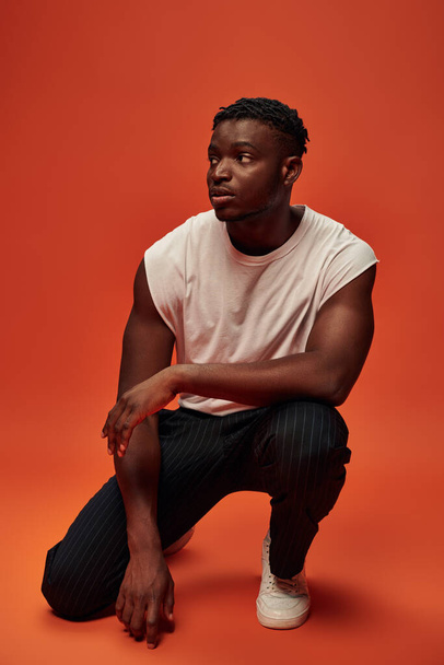 serious african american man in white tank top sitting on haunches and looking away on red backdrop - Zdjęcie, obraz