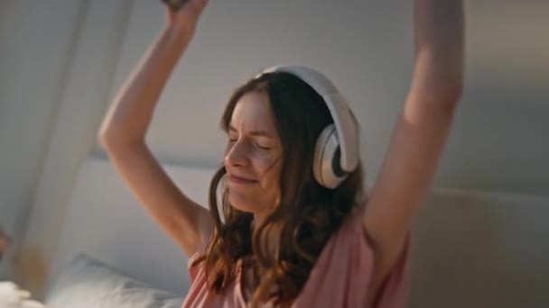 Šťastná holka tančí sluchátka doma. Mladá žena poslouchající hudbu drží smartphone. Energická radostná fena si užívá oblíbenou píseň melodie. Uvolněný usměvavý model při výběru seznamu skladeb - Záběry, video