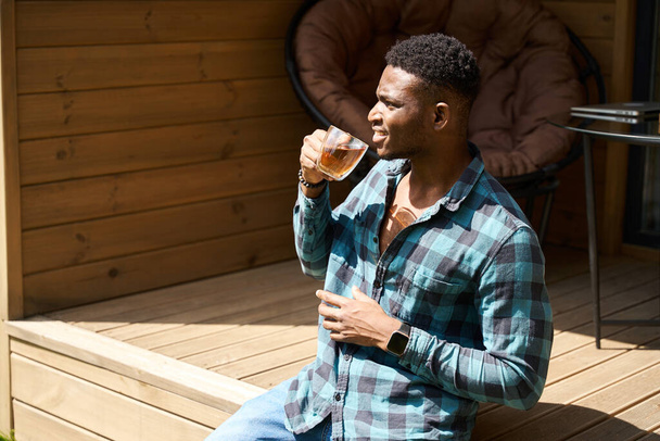 Afroameričan na dřevěné terase s šálkem čaje, užívá si letní dopoledne - Fotografie, Obrázek