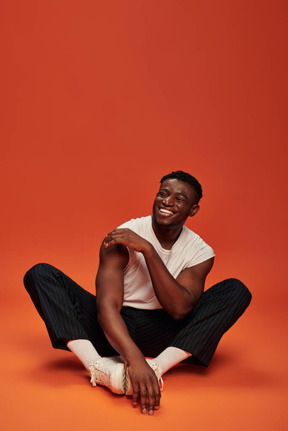 emocionado hombre afroamericano elegante sentado y riendo con los ojos cerrados, fondo rojo y naranja - Foto, imagen