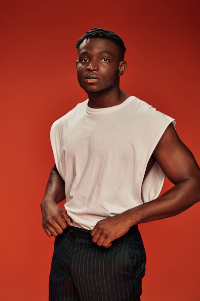 pensive african american man in white tank top and black pants looking away on red backdrop - Photo, Image