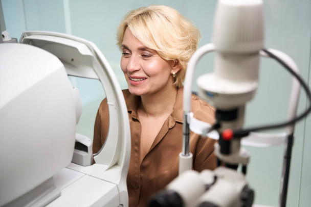 Усміхнена жінка сидить перед спеціальним офтальмологічним пристроєм, жінка за зустріччю зі спеціалістом - Фото, зображення