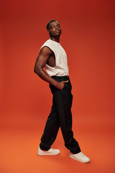 self-confident african american man in white tank top with hand in pocket of black pants on red - Фото, зображення