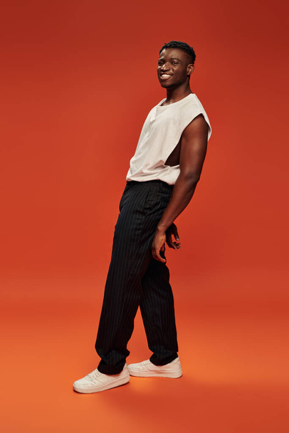 despreocupado hombre afroamericano en pantalones negros y camiseta blanca posando sobre fondo rojo y naranja - Foto, imagen