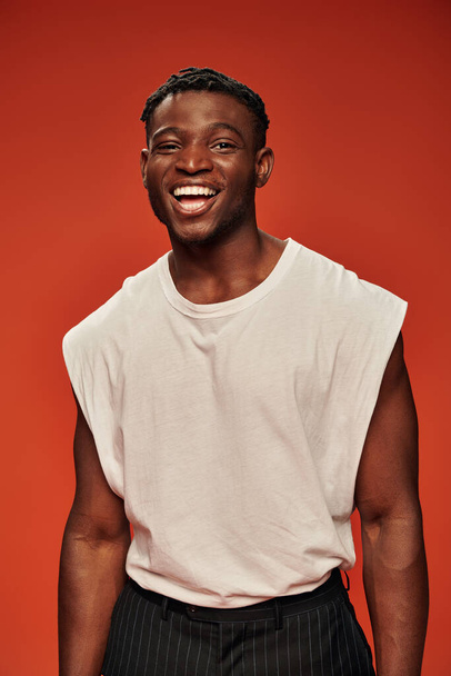 young excited african american male model in white tank top laughing and looking at camera on red - Foto, afbeelding