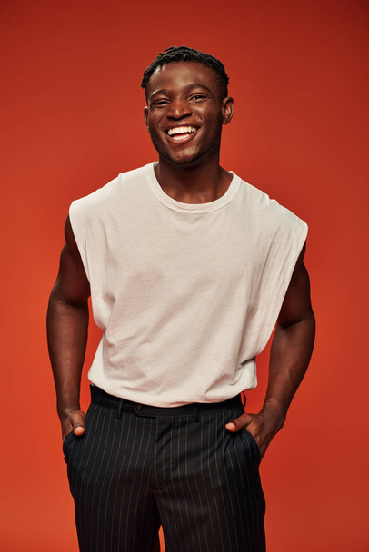 Веселый африканский американец с руками в карманах улыбается в камеру на красного, горячего и счастливого парня - Фото, изображение