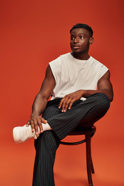 caldo ragazzo americano africano in pantaloni neri e canotta bianca in posa sulla sedia e guardando lontano sul rosso - Foto, immagini