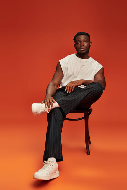 african american male model in stylish street wear sitting on chair on red and orange backdrop - Фото, изображение