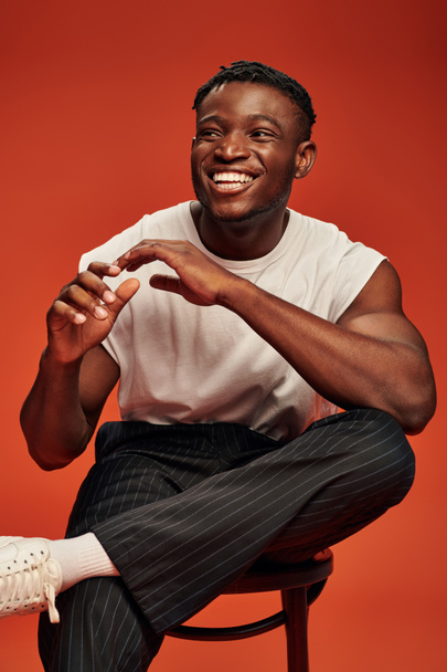 laughing african american man in white tank top sitting on chair and gesturing on red backdrop - Фото, изображение