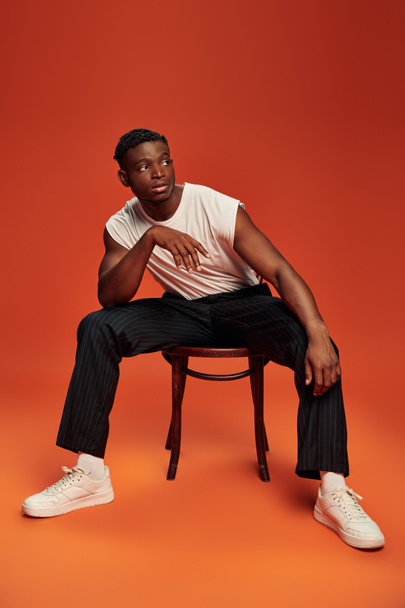 uomo afroamericano moderno ed elegante seduto sulla sedia e guardando lontano sullo sfondo rosso e arancione - Foto, immagini