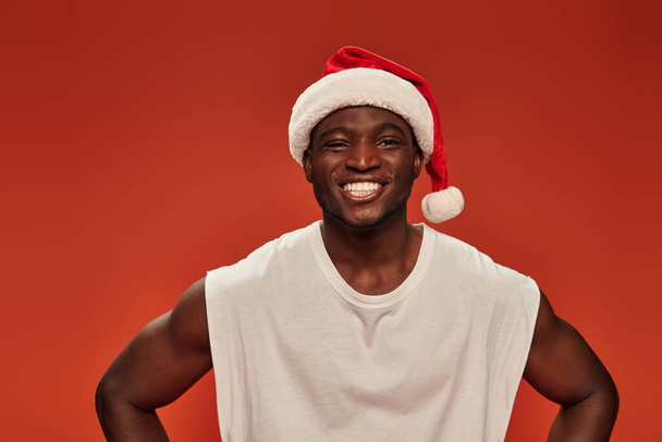 homme afro-américain joyeux avec un sourire radieux posant dans le chapeau du Père Noël et regardant la caméra sur rouge - Photo, image