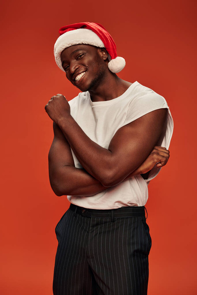 horký a atletický africký Američan chlap v Santa klobouku a bílé tílko s úsměvem na kameru na červené - Fotografie, Obrázek