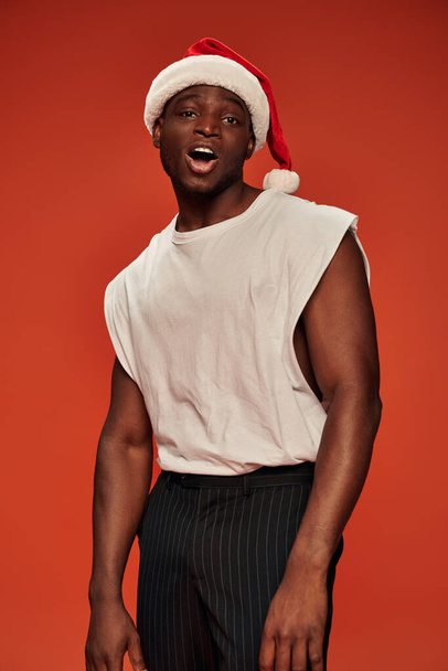 flirty african american man in santa hat and white tank looking at camera on red backdrop - Photo, image